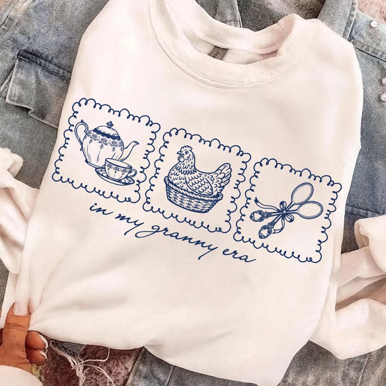 May include: White sweatshirt with a blue graphic design featuring a teapot and teacup, a hen in a basket, and kitchen scissors. The phrase "in my granny era" is written below the graphics.