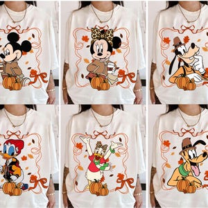 Puede incluir: Camisetas color crema con varios personajes de Disney con atuendos otoñales. Cada camiseta muestra un personaje diferente, incluyendo Mickey Mouse, Minnie Mouse, Goofy, Donald Duck, Daisy Duck y Pluto, con calabazas y hojas de otoño. Los diseños están enmarcados con un borde decorativo.