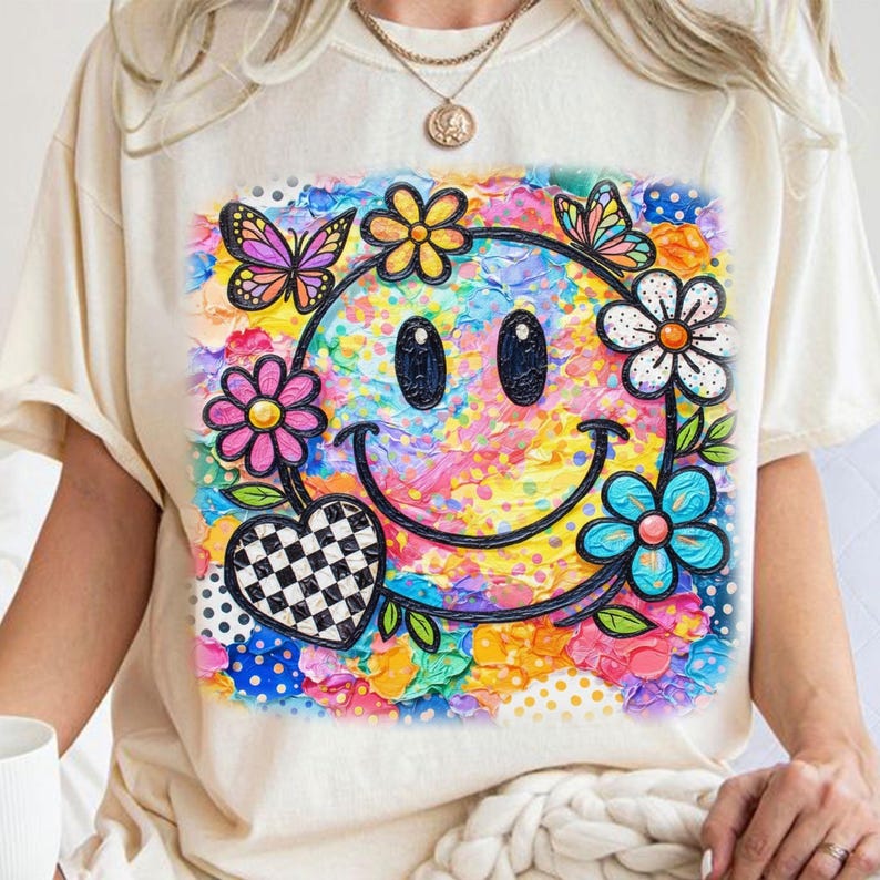 以下が含まれることがあります： カラフルなスマイリーフェイスのデザインが施された、クリーム色のTシャツ。グラフィックには、花、蝶、市松模様のハートで囲まれた、スマイリーフェイスの黒いアウトラインが含まれています。背景は明るい色のミックスです。