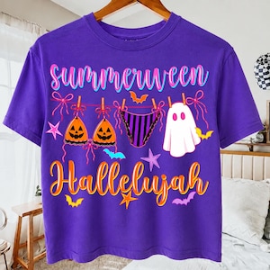 May include: A purple short-sleeve t-shirt featuring a graphic print of a clothesline with two jack-o'-lantern bikini tops, striped bikini bottoms, and a ghost. The text reads "summerween" and "Hallelujah" in orange and pink script lettering.