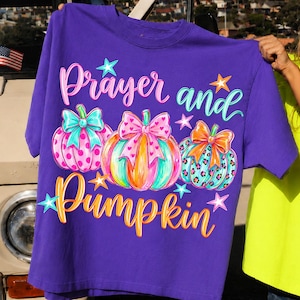 May include: A purple t-shirt featuring the words "Prayer and Pumpkin" in a whimsical font. The shirt design includes three colorful pumpkins with bows and star accents. The pumpkins have polka dots, hearts, and leopard print patterns.