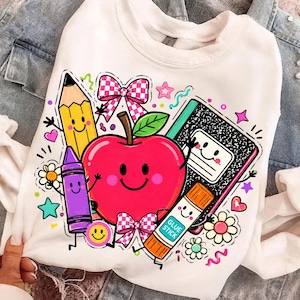May include: A white sweatshirt featuring a colorful graphic print of a smiling red apple surrounded by school supplies including a yellow pencil, a purple crayon, a black and white composition notebook, and an orange glue stick with text.
