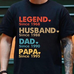 May include: Black t-shirt with the words "LEGEND Since 1968", "HUSBAND Since 1988", "DAD Since 1990", and "PAPA Since 1995" in various colors. The shirt has a crew neck and short sleeves.