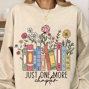 May include: Beige sweatshirt with a graphic of colorful books and flowers. The text "Just one more chapter" is printed below the graphic.