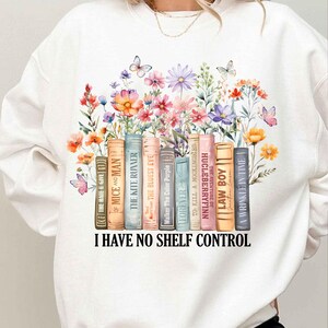 May include: White sweatshirt with a colorful floral design and a stack of books with titles like "The Kite Runner", "To Kill a Mockingbird", and "The Bluest Eye". The text "I Have No Shelf Control" is printed below the books.