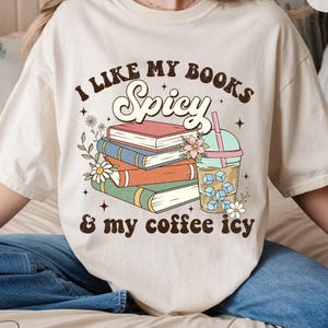 May include: A beige t-shirt with the text "I like my books spicy & my coffee icy" in brown lettering. The text is surrounded by a stack of books, a cup of iced coffee, and floral illustrations.