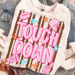 May include: A white crewneck sweatshirt featuring a graphic print with the text TOUCH DOWN in large pink letters. The design includes brown football-textured stripes, a pink football, a pink helmet, a pink megaphone with GO TEAM, and stars.