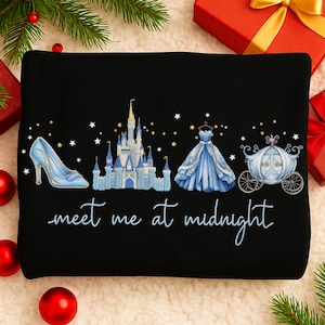 Puede incluir: Sudadera negra con temática de Cenicienta. Presenta un zapato de cristal azul, un castillo, un vestido de baile y un carruaje. El texto "meet me at midnight" está escrito en cursiva. Decoraciones festivas de vacaciones en el fondo.