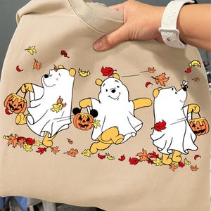 May include: Beige sweatshirt featuring a Halloween-themed design. The graphic depicts three cartoon bears dressed as ghosts, each holding a jack-o'-lantern. Autumn leaves are scattered around the bears. The sweatshirt is held by a person wearing a white watch.