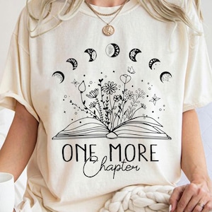 May include: A white t-shirt with a black graphic design of a book with flowers and moons growing out of it. The text "One More Chapter" is printed below the book.