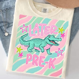 Peut inclure: T-shirt cr&egrave;me avec une illustration d'alligator turquoise et le texte rose "LATER PRE-K". Le motif comprend des &eacute;toiles roses et turquoise et un fond ray&eacute; rose, turquoise et blanc.