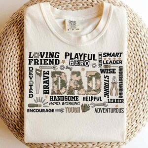 Peut inclure: T-shirt crème avec le mot "DAD" en imprimé camouflage. Autour du mot se trouvent d'autres mots comme "Loving", "Playful", "Hero" et "Brave". Le t-shirt porte également le logo Comfort Colors.
