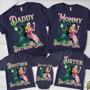 Puede incluir: Camisetas azul marino con diseños temáticos de El Mago de Oz. Los diseños incluyen "Daddy", "Mommy", "Brother" y "Sister" con ilustraciones de la Bruja Malvada y Dorothy. Un body dice "Dorothy".