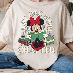 Op de afbeelding: Een wit T-shirt met een vintage-stijl afbeelding van Minnie Mouse die een boek leest. De tekst "Just One More Chapter" is in een cirkel rond Minnie Mouse gedrukt.