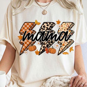May include: Cream-colored t-shirt featuring the word "mama" in a black script, overlaid on three lightning bolt designs. The lightning bolts have fall-themed patterns, including orange plaid, leopard print, and floral designs, with pumpkins and leaves.