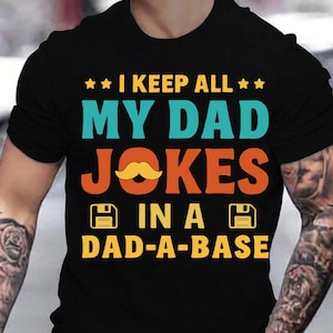 May include: Black t-shirt with a retro-style design. The text reads "I keep all my dad jokes in a dad-a-base" with a floppy disk icon on either side of the text. A yellow moustache is above the word "jokes".