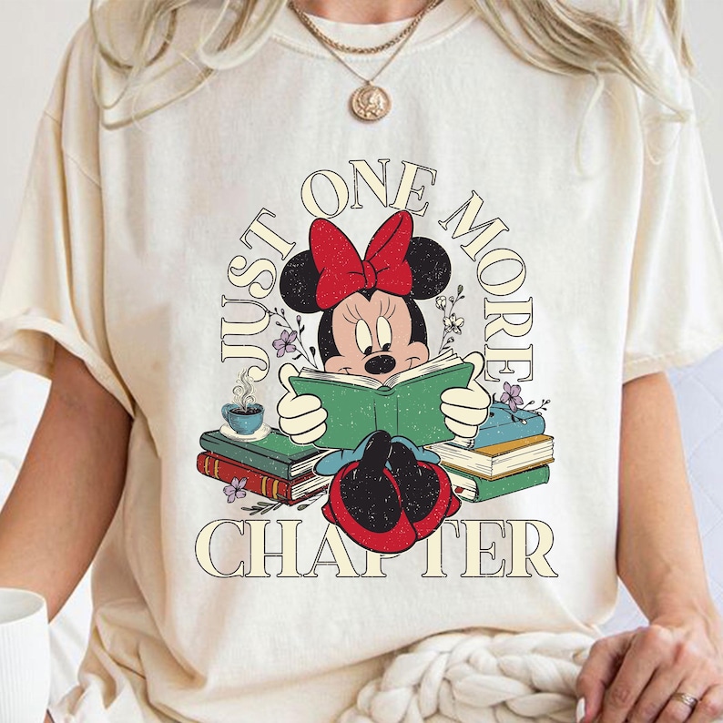 Op de afbeelding: Een wit T-shirt met een cartoon afbeelding van Minnie Mouse die een boek leest. De tekst "Just One More Chapter" is rond de afbeelding gedrukt.