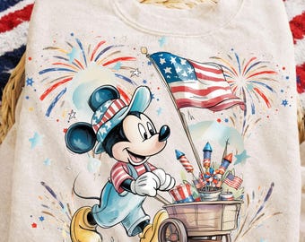 May include: A beige sweatshirt with a patriotic Mickey Mouse design. Mickey is shown pushing a cart of fireworks and holding an American flag. The design incorporates fireworks, stars, and stripes in red, white, and blue.