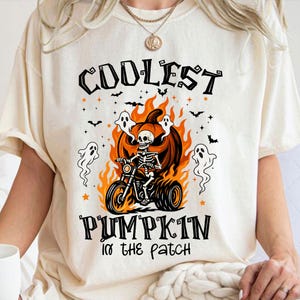 Può includere: T-shirt color crema con grafica a tema Halloween. Il design presenta uno scheletro su una moto davanti a una zucca in fiamme, con le parole "COOLEST PUMPKIN IN THE PATCH" in nero. Sono inclusi anche fantasmi e pipistrelli.