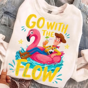 May include: A white crewneck sweatshirt featuring a graphic of a cartoon cowboy character wearing a brown hat and yellow shirt, lounging on a pink flamingo pool float in blue water, with the yellow text GO WITH THE FLOW and small stars.