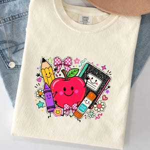 May include: A cream-colored t-shirt featuring a graphic print of a smiling red apple surrounded by a yellow pencil, a purple crayon, a black composition notebook, and a glue stick, all with faces, accented by pink checkered bows and stars.