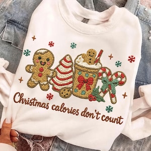 May include: White sweatshirt with embroidered Christmas-themed design. Features a gingerbread man, Christmas tree, coffee, candy cane, and the text "Christmas calories don't count" in brown thread. Accented with snowflakes and cross shapes.