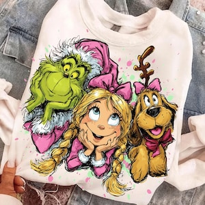 Könnte beinhalten: Weißes Sweatshirt mit einer farbenfrohen Grafik von Grinch, Cindy Lou Who und Max dem Hund. Der Grinch ist grün, Cindy Lou Who hat blonde Zöpfe und Max ist braun mit Geweih. Rosa Akzente und Farbspritzer vervollständigen das Design.