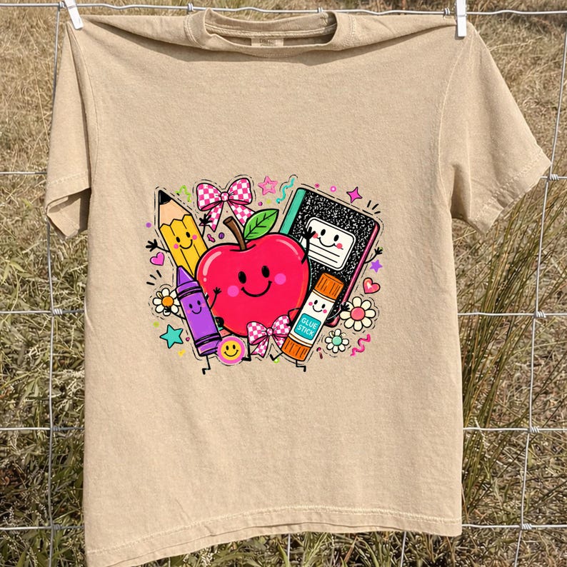 May include: A tan t-shirt featuring a colorful graphic print of a smiling red apple surrounded by school supplies including a yellow pencil, a purple crayon, a black and white composition notebook, and an orange glue stick labeled GLUE STICK.
