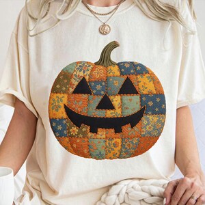 May include: Cream-colored t-shirt featuring a patchwork pumpkin design. The pumpkin is composed of various fabric squares in orange, blue, and yellow floral patterns, with a black face and a green stem. A festive, seasonal graphic tee.