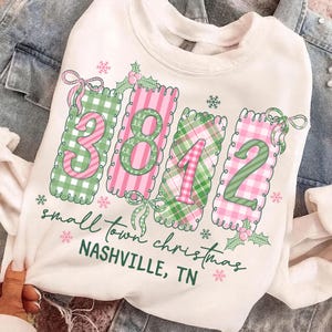 Könnte beinhalten: Weißes Sweatshirt mit weihnachtlichem Design. Das Design zeigt die Zahlen "3812" in rosa und grün kariert, mit den Worten "small town christmas" und "NASHVILLE, TN" in grüner Schrift. Schneeflocken und Stechpalmenakzente vervollständigen den festlichen Look.