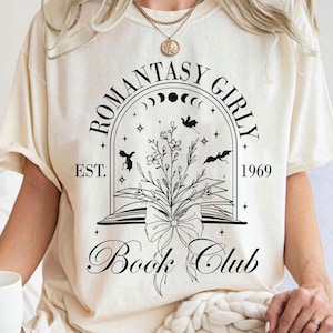 May include: Cream-colored t-shirt with a black graphic design. The design features the words "ROMANTASY GIRLY" arched above a floral and bat illustration, with "Book Club" below. The year "1969" is also included.