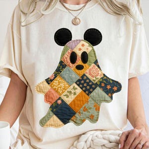 May include: Cream-colored t-shirt featuring a patchwork ghost design with Mickey Mouse ears. The ghost is made of various floral and geometric patterned fabric squares in shades of orange, blue, green, and yellow. Black oval eyes and mouth complete the design.