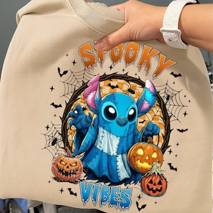 May include: Beige sweatshirt featuring a blue cartoon character with pink ears and a blue body, holding a jack-o'-lantern. The words "Spooky Vibes" are printed in orange and blue. The design includes bats, spiderwebs, and pumpkins.