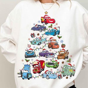 Könnte beinhalten: Weißes Sweatshirt mit einem Weihnachtsbaum-Design aus Zeichentrickautos mit Weihnachtsmützen. Die Autos sind in verschiedenen Farben gehalten, und der Baum ist mit Lichtern, Schneeflocken und Geschenkboxen geschmückt. Ein goldener Stern ziert die Spitze des Baumes.