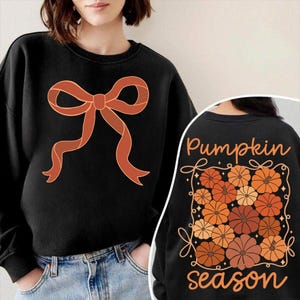 May include: Black sweatshirt featuring a large orange bow design on the front. The back of the sweatshirt displays the text "Pumpkin season" with a pumpkin graphic in shades of orange and brown.