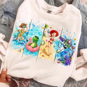 Könnte beinhalten: Weißes Sweatshirt mit einem farbenfrohen Strandmotiv. Das Design zeigt Cartoon-Charaktere, darunter ein Mann mit Strohhut, ein grüner Dinosaurier auf einem rosa Schwimmer, ein Mädchen in einem roten Badeanzug und Buzz Lightyear.