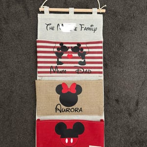 May include: A fabric wall organiser with four pockets, a wooden dowel, and a hanging string. The pockets feature Disney-themed designs, including Mickey and Minnie Mouse silhouettes, names, and the text "The Family" at the top.