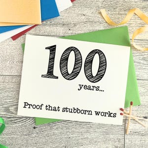 May include: A white greeting card with the number "100" in large, black, handwritten-style font. The text "years..." and "Proof that stubborn works" are below. The card is on a green envelope, with matches and ribbon nearby.