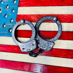May include: Ornate, engraved handcuffs with a dark finish, connected by a chain. The handcuffs are set against a backdrop of a wooden American flag with red, white, and blue stripes and stars.