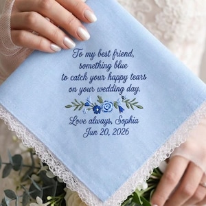 Op de afbeelding: Een lichtblauwe vierkante zakdoek met witte kanten rand en donkerblauw borduurwerk met de tekst: To my best friend, something blue to catch your happy tears on your wedding day. Love always, Sophia Jun 20, 2026, en een bloemmotief in het midden.