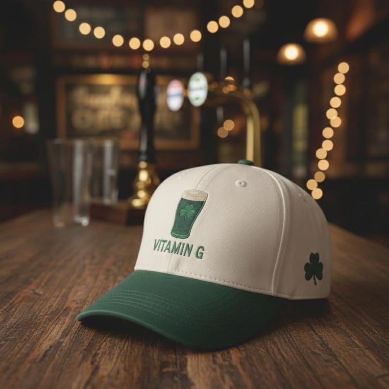 May include: A white and green baseball cap with a shamrock and the words "Vitamin G." The cap features a green brim and a design of a green beer glass with a shamrock. A small shamrock is on the side.