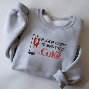 Puede incluir: Una sudadera gris claro con el texto "IN CASE OF ACCIDENT, MY BLOOD TYPE IS Diet Coke" en rojo y blanco. El diseño incluye un gráfico de bolsa de sangre Diet Coke. La sudadera está doblada sobre una superficie blanca.