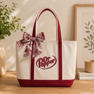 May include: A cream-colored canvas tote bag featuring the embroidered red Dr Pepper logo. The bag has deep red trim, two matching handles, and a decorative bow tied to one handle, patterned with a red and white toile-style print.