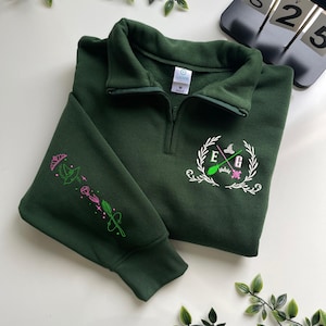 May include: Dark green sweatshirt with a quarter-zip collar. The left sleeve features embroidered pink and green magical elements. The chest displays a white embroidered crest with the letters E and G, a witch's hat, a broom, and a crown.
