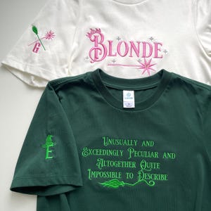 Puede incluir: Dos camisetas, una blanca con bordado rosa que dice "BLONDE" y la otra verde con bordado verde que dice "UNUSUALLY AND EXCEEDINGLY PECULIAR AND ALTOGETHER QUITE IMPOSSIBLE TO DESCRIBE".