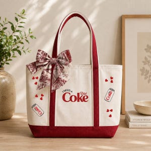 Pode incluir: Uma bolsa tote de lona na cor creme com acabamento e alças em vermelho escuro. A frente apresenta o texto "I NEED A Diet Coke" em vermelho, rodeado por pequenos ícones de corações vermelhos e duas ilustrações de latas de Diet Coke.
