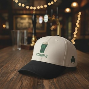 May include: A white and black baseball cap with a green shamrock and a green beer glass design. The text "VITAMIN G" is printed on the front. A green shamrock is on the side. The cap is on a wooden surface.