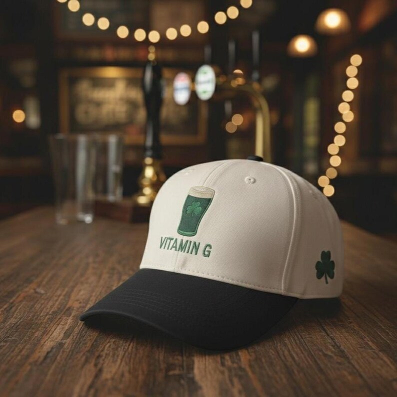 May include: A white and black baseball cap with a green shamrock and a green beer glass design. The text "VITAMIN G" is printed on the front. A green shamrock is on the side. The cap is on a wooden surface.