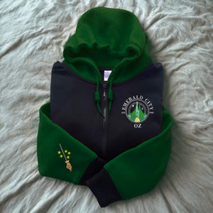 May include: A black and green zip-up hoodie featuring the words "Emerald City OZ" and a castle graphic. The sleeves have green cuffs and a gold embroidered witch's broom with yellow stars. The hood is green.