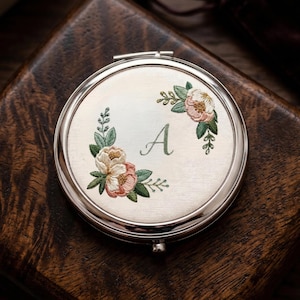 Puede incluir: Un espejo compacto redondo y plateado con un diseño floral bordado. El diseño presenta flores rosas y crema con hojas verdes, que rodean la letra "A". El espejo está sobre una superficie de madera oscura.